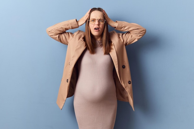 Foto mulher grávida chocada e desesperada com um belo vestido e casaco posando isolada sobre um fundo azul fazendo caretas de sons altos ouvindo barulhos desagradáveis