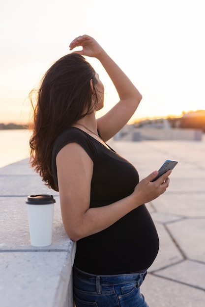 Mulher grávida com cabelo comprido mantém o smartphone ao ar livre. Vista lateral. Dia caloroso. Nova vida, novos horizontes e conceito de oportunidades