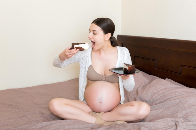 Mulher grávida com fome está comendo um pedaço de bolo saboroso relaxando na cama em casa Doces desejos durante a gravidez