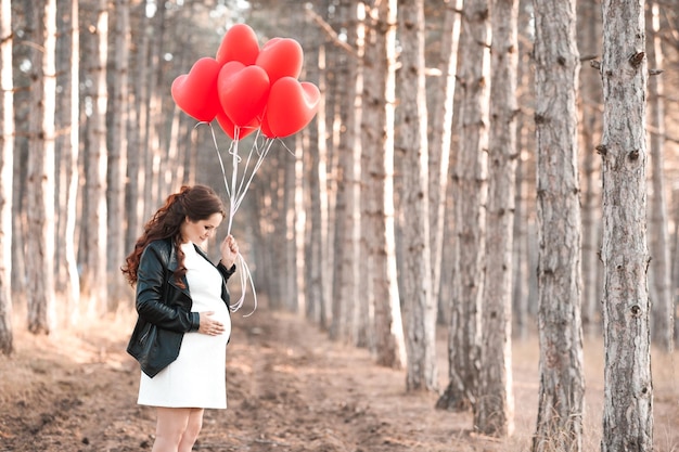 Mulher grávida de 30 a 35 anos segurando balões vermelhos em forma de coração em pé na floresta