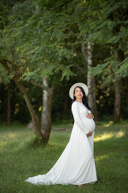 Mulher grávida de chapéu posando em um vestido branco sobre um fundo de árvores verdes
