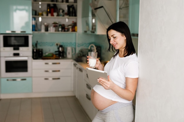 Mulher grávida de meia idade usando tablet digital enquanto toma leite na cozinha em casa