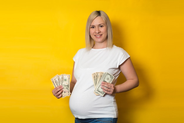 Foto mulher gravida de sorriso feliz com dinheiro, dólares em uma parede amarela. benefícios para mulheres grávidas