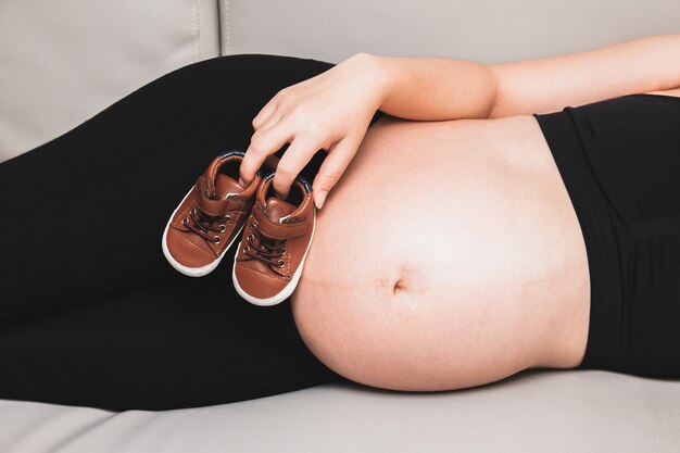 Mulher grávida dorme no sofá com a mão segurando sapatinhos de bebê perto da barriga