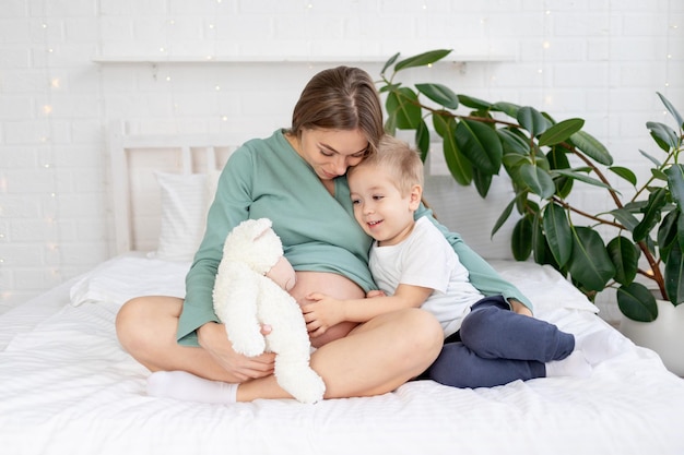 Mulher grávida e filho bebê tocando barrigão em casa e acariciando na cama, o conceito de gravidez e a espera do nascimento de um bebê e um segundo filho na família
