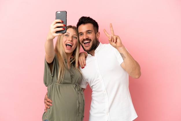 Mulher grávida e homem sobre fundo rosa isolado fazendo uma selfie com o celular