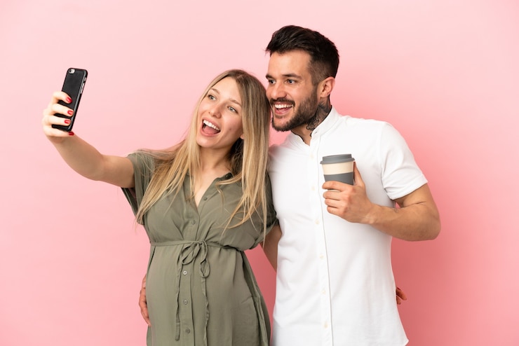 Foto mulher grávida e homem sobre fundo rosa isolado fazendo uma selfie com o celular