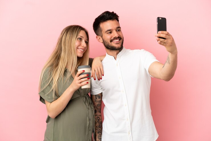 Foto mulher grávida e homem sobre fundo rosa isolado fazendo uma selfie com o celular