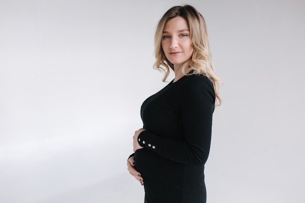 Mulher grávida elegante em um vestido preto com as mãos na barriga e um sorriso no fundo branco