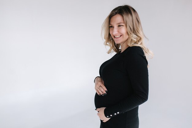 Mulher grávida elegante em um vestido preto com as mãos na barriga e um sorriso no fundo branco