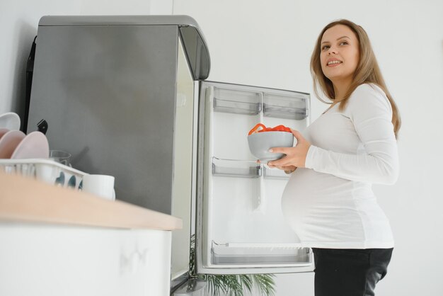 Mulher grávida em casa na cozinha abre a geladeira