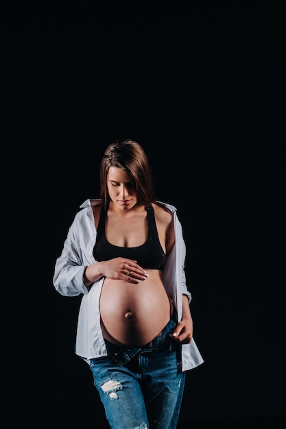 Mulher grávida em jeans e camisa branca em estúdio em fundo preto.