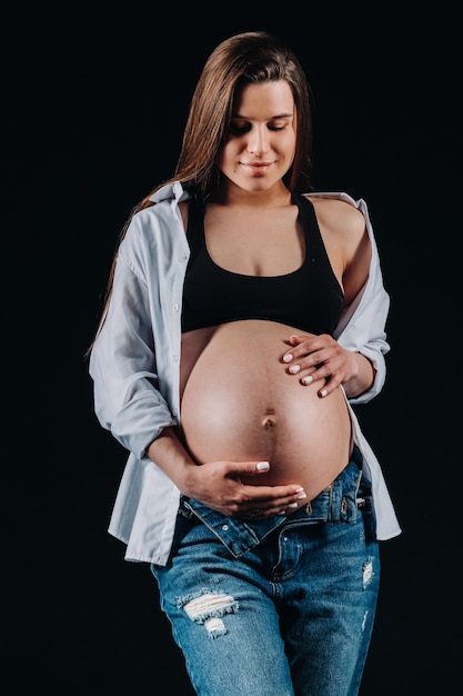 Mulher grávida em jeans e camisa branca em estúdio em fundo preto.