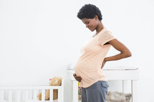 Mulher grávida em pé perto do berço e sorrindo enquanto olha para o estômago