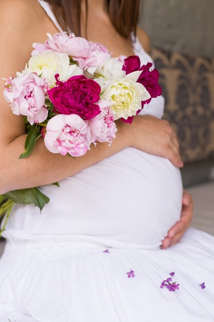 mulher grávida em um lindo vestido branco tocando a barriga com as mãos e segurando um buquê de peônias