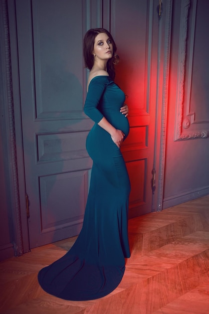 Mulher grávida em um longo vestido azul de pé contra a parede no estúdio
