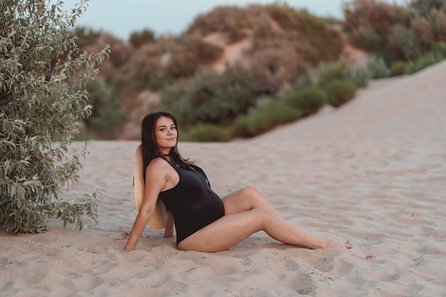 mulher grávida em um maiô preto e chapéu em uma praia de areia