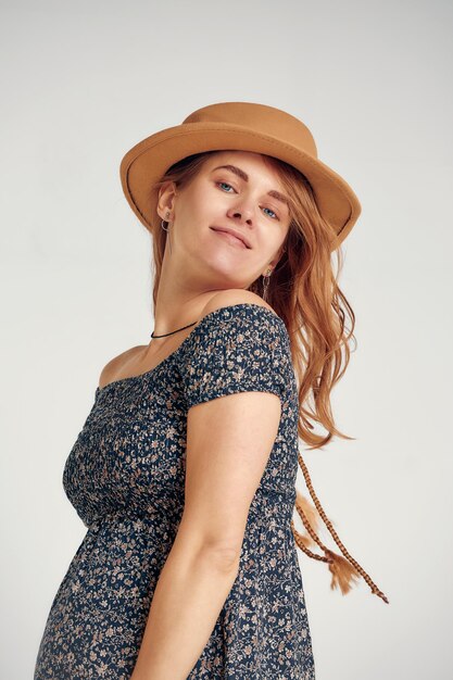 Foto mulher grávida em vestido de verão de estilo country e chapéu de palha de pé tocando sua barriga estúdio