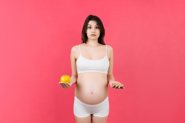 mulher grávida fazendo uma escolha entre uma maçã e uma pilha de pílulas em fundo colorido com espaço de cópia Tomando vitaminas durante a gravidez conceito