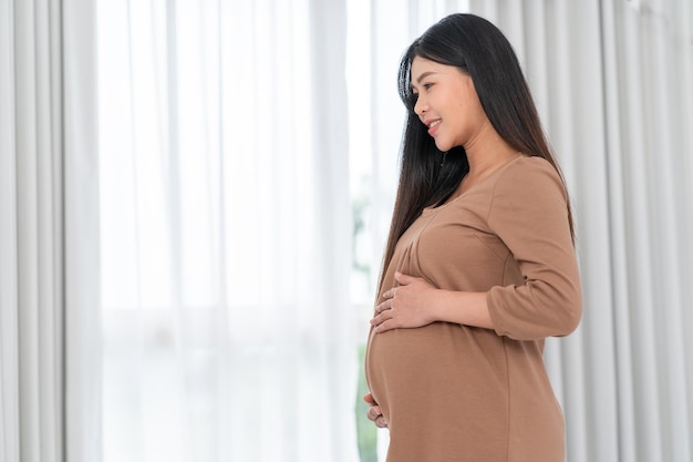 Mulher grávida feliz asiática está de pé ao lado de janelas e tocando sua barriga, gravidez, maternidade, pessoas e conceito de expectativa