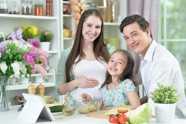 Mulher grávida feliz com marido e filha preparando salada juntos na cozinha