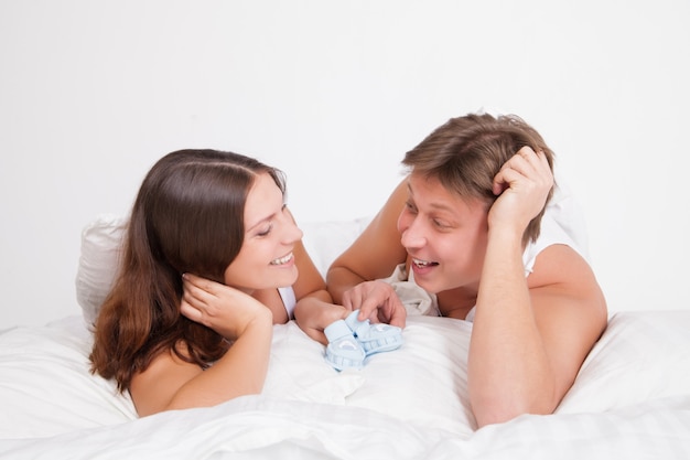 Mulher grávida feliz com o marido na cama branca em um quarto claro com botinhas de bebê