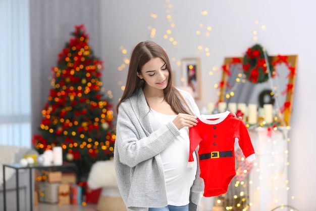 Mulher grávida feliz com roupa de bebê de papai noel em quarto decorado para o natal