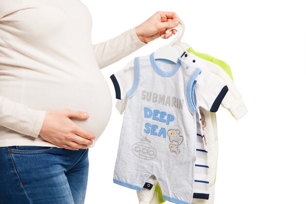 Mulher grávida feliz segurando diferentes peças de roupa infantil sobre fundo branco