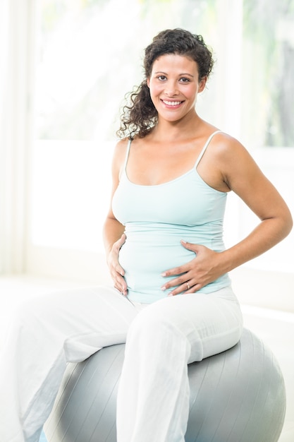 Mulher grávida feliz tocando sua barriga enquanto está sentada na bola de exercício