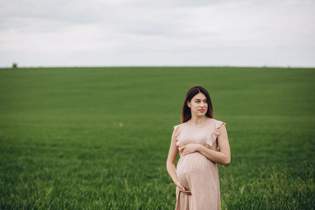 Mulher grávida fica no campo com grama verde e abraça sua barriga Vibrações de verão da luz do sol