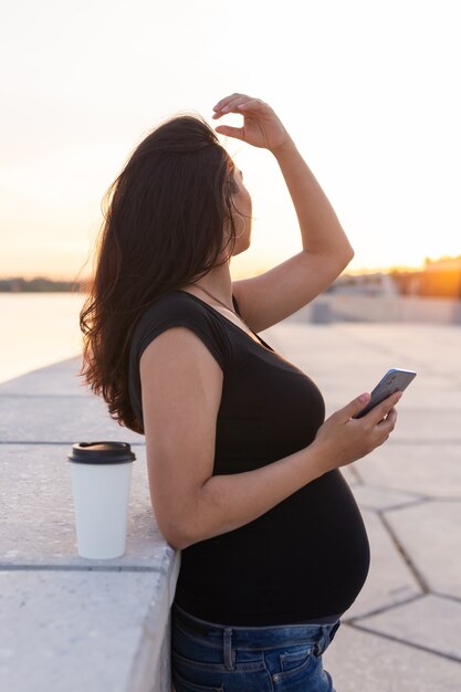 Mulher grávida hispânica segurando um smartphone e relaxando em um aterro com luz quente pela manhã