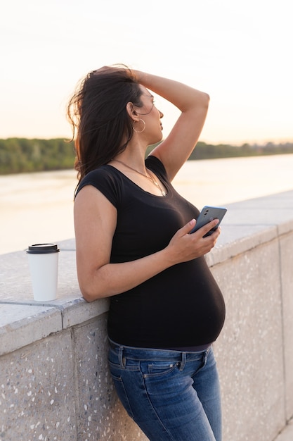 Mulher grávida hispânica segurando um smartphone e relaxando em um aterro com luz quente pela manhã