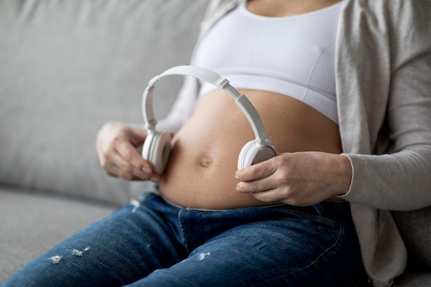 Mulher grávida irreconhecível segurando fones de ouvido sem fio perto da barriga tocando música para o bebê