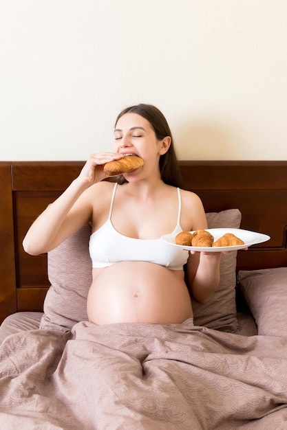 Foto mulher grávida jovem com café da manhã de leite e croissant sentada na cama e olhando para a barriga