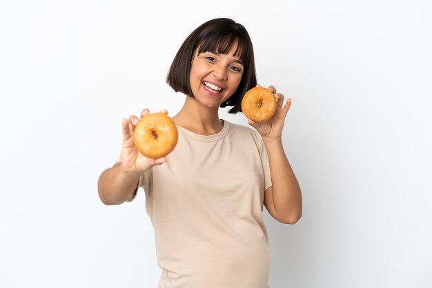 Mulher grávida jovem mestiça isolada no fundo branco segurando rosquinhas com uma expressão feliz