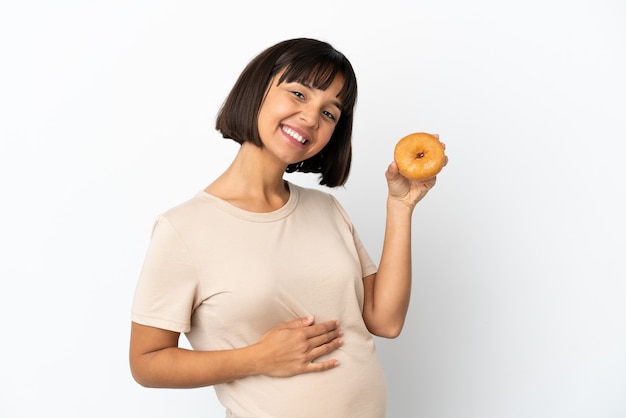 Mulher grávida jovem mestiça isolada no fundo branco segurando uma rosquinha feliz