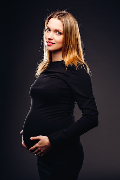 Mulher gravida macia e sensual bonita no vestido preto que está contra um fundo da parede cinzenta no estúdio.