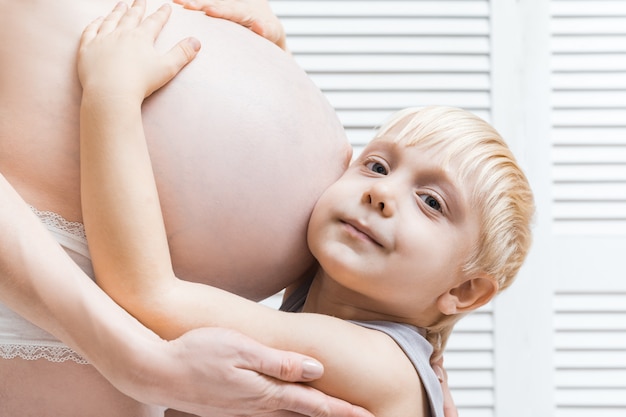 Mulher grávida mãe e filho estão abraçando. Garoto bonito inclinou a orelha para a barriga da mãe