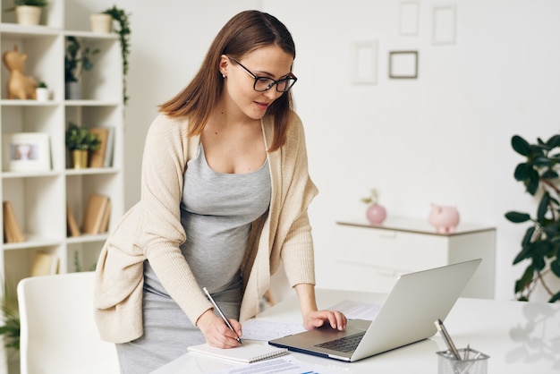 Mulher grávida moderna usando óculos e casaco de lã em pé à mesa e usando o laptop enquanto escreve tarefas no bloco de notas