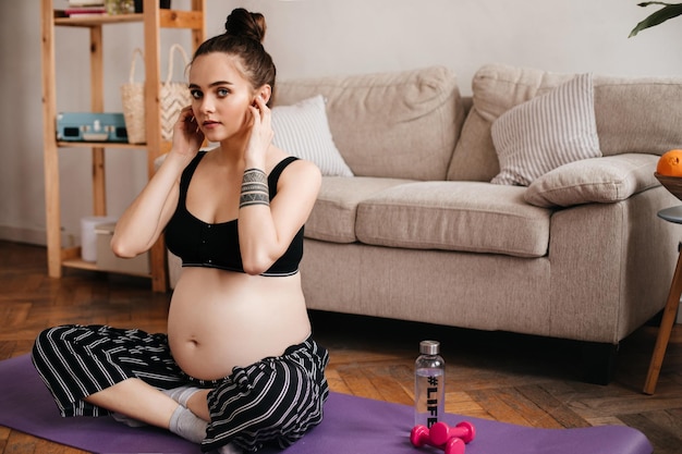 Mulher grávida morena de olhos azuis em top cortado preto e calças listradas olha para a câmera na sala de estar garota atraente sentada no tapete de ioga