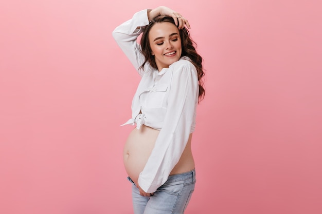 Mulher grávida morena feliz em calças jeans e camisa branca cortada sorri amplamente em poses de garota encaracolada isolada em fundo rosa