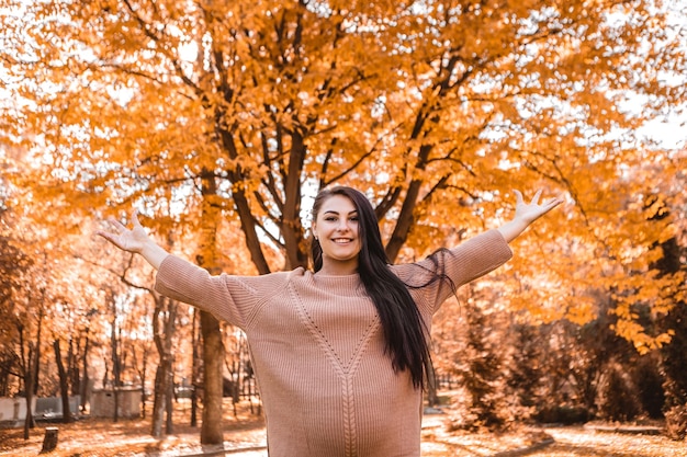 Mulher grávida parada na floresta do parque da cidade do outono, levantando as mãos, barriga redonda com bebê