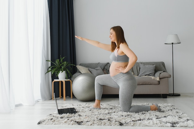 Mulher grávida praticando ioga em casa com o laptop. Mãe grávida fazendo aula de treinamento em vídeo pré-natal dentro de casa. Exercícios femininos, medite durante a gravidez. Aula de fitness online em dispositivos digitais.