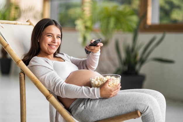 Mulher grávida relaxando na cadeira em casa assistindo TV e comendo pipoca
