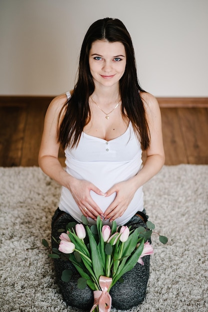 Mulher grávida segurando a barriga com um buquê de flores abraçando a barriga fazendo símbolo coração mãos em casa Chá de bebê Conceito de maternidade Menina na foto