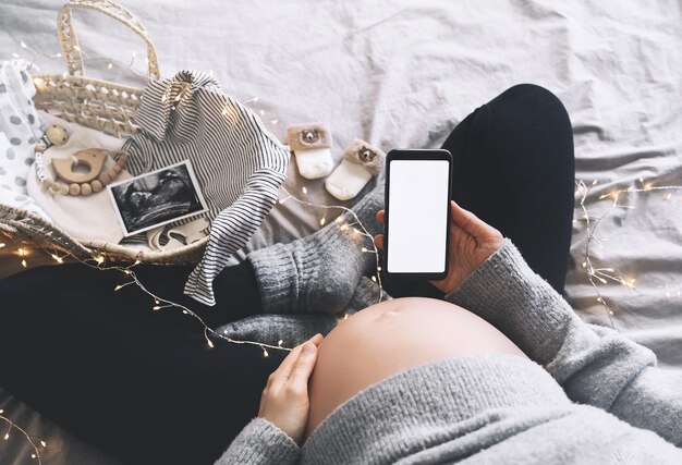 Mulher grávida segurando nas mãos um smartphone com visor branco vazio