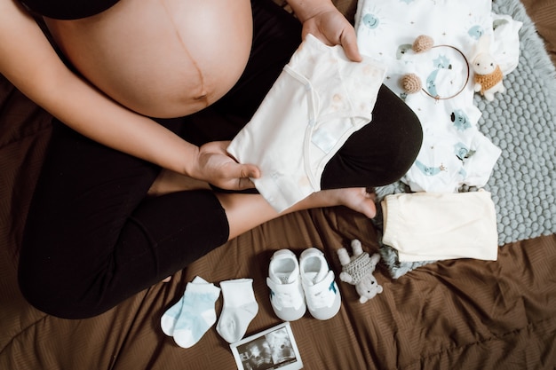 Mulher grávida segurando o body do bebê no quarto com um brinquedo e as necessidades para o parto recém-nascido. mulher grávida esperando e se preparando para o nascimento do bebê. conceito de mulher grávida.