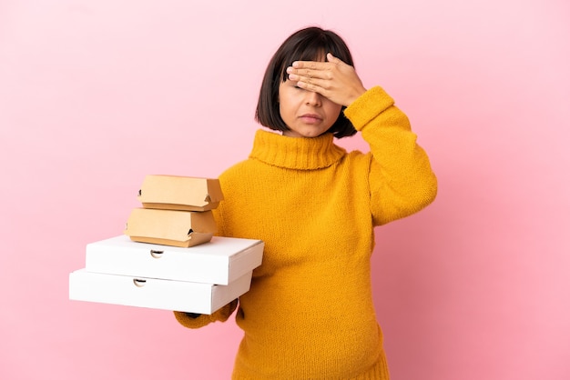 Mulher grávida segurando pizzas e hambúrgueres isolados no fundo rosa, cobrindo os olhos com as mãos. não quero ver nada