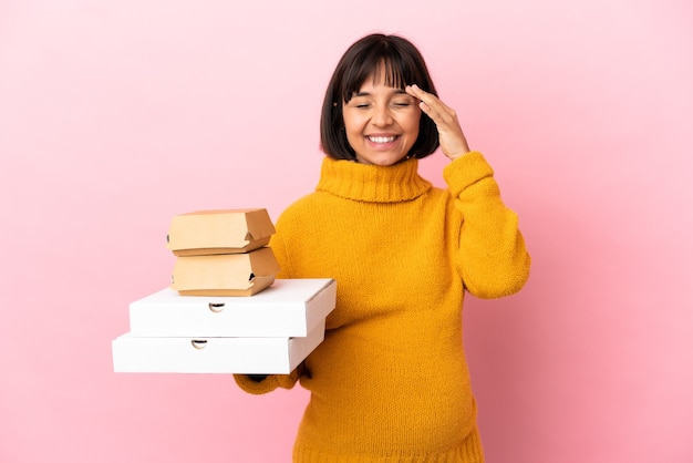 Mulher grávida segurando pizzas e hambúrgueres isolados no fundo rosa e sorrindo muito