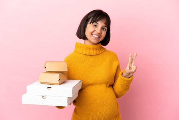 Mulher grávida segurando pizzas e hambúrgueres isolados no fundo rosa, sorrindo e mostrando o sinal da vitória